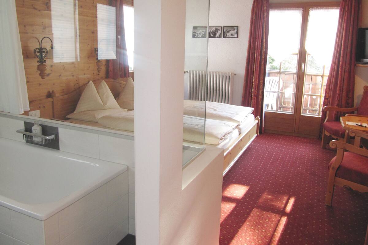 A hotel room with a bed, a chair, a small table, a radiator, and a window with curtains.