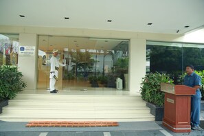 Entrada de un hotel con una zona de recepción, un hombre de pie frente a un atril y un guardia de seguridad.