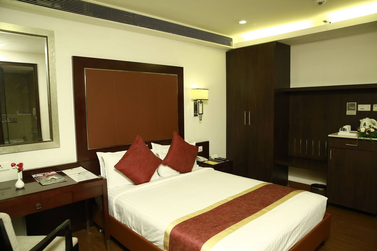 A hotel room with a large bed, a desk with a chair, a nightstand, a wardrobe, and a mirror.