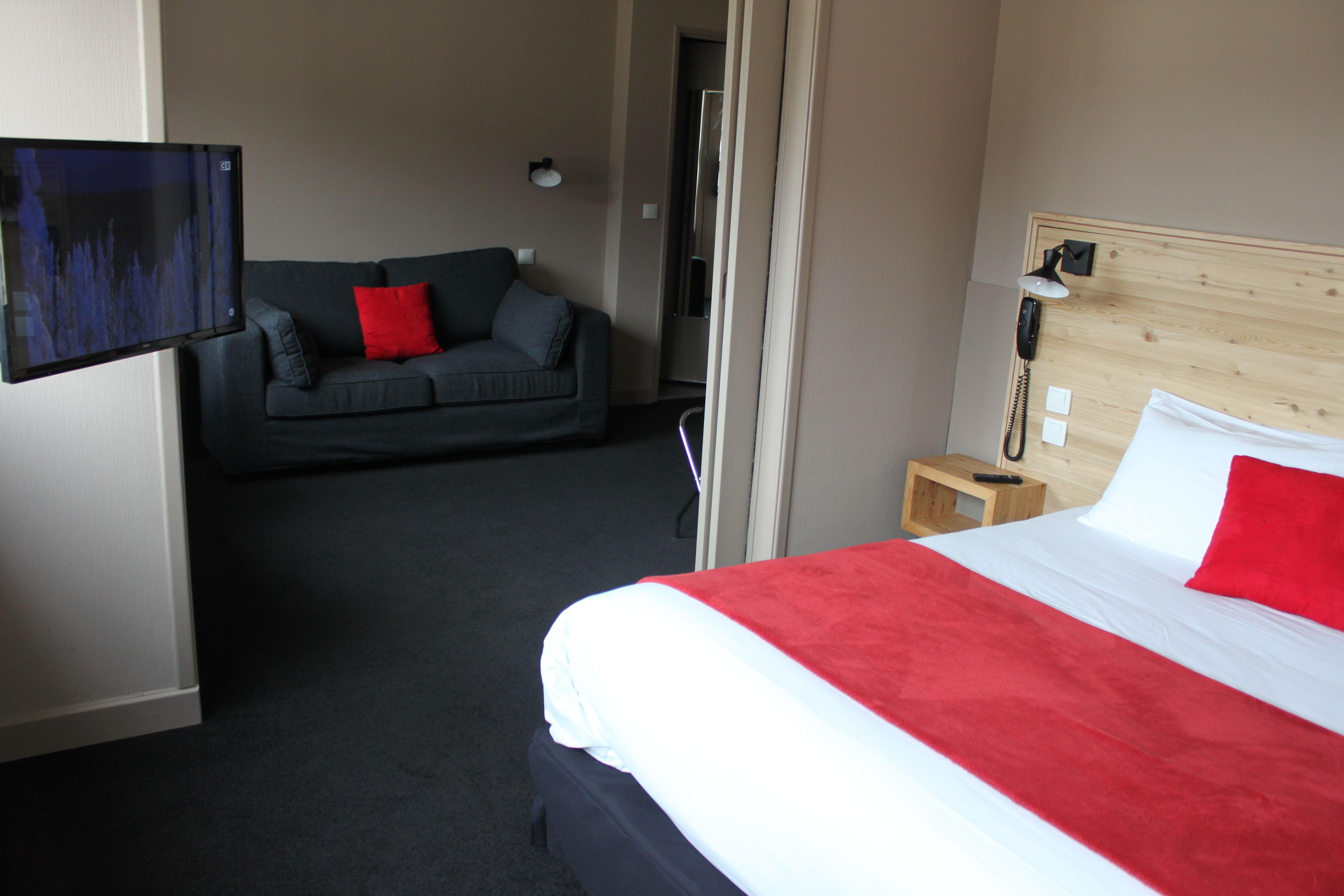 Habitación de hotel con cama, sofá, televisión, mesita de noche y cabecera de madera.