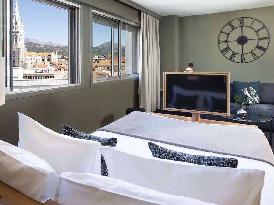 A hotel room with a bed, a TV, a sofa, and a large window offering a cityscape view.