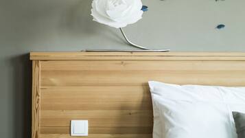 Una cama bien hecha con ropa blanca, una mesita de noche de madera con una tetera blanca y una taza, y una pared decorada con una gran flor de papel blanca y adornos de mariposas azules.