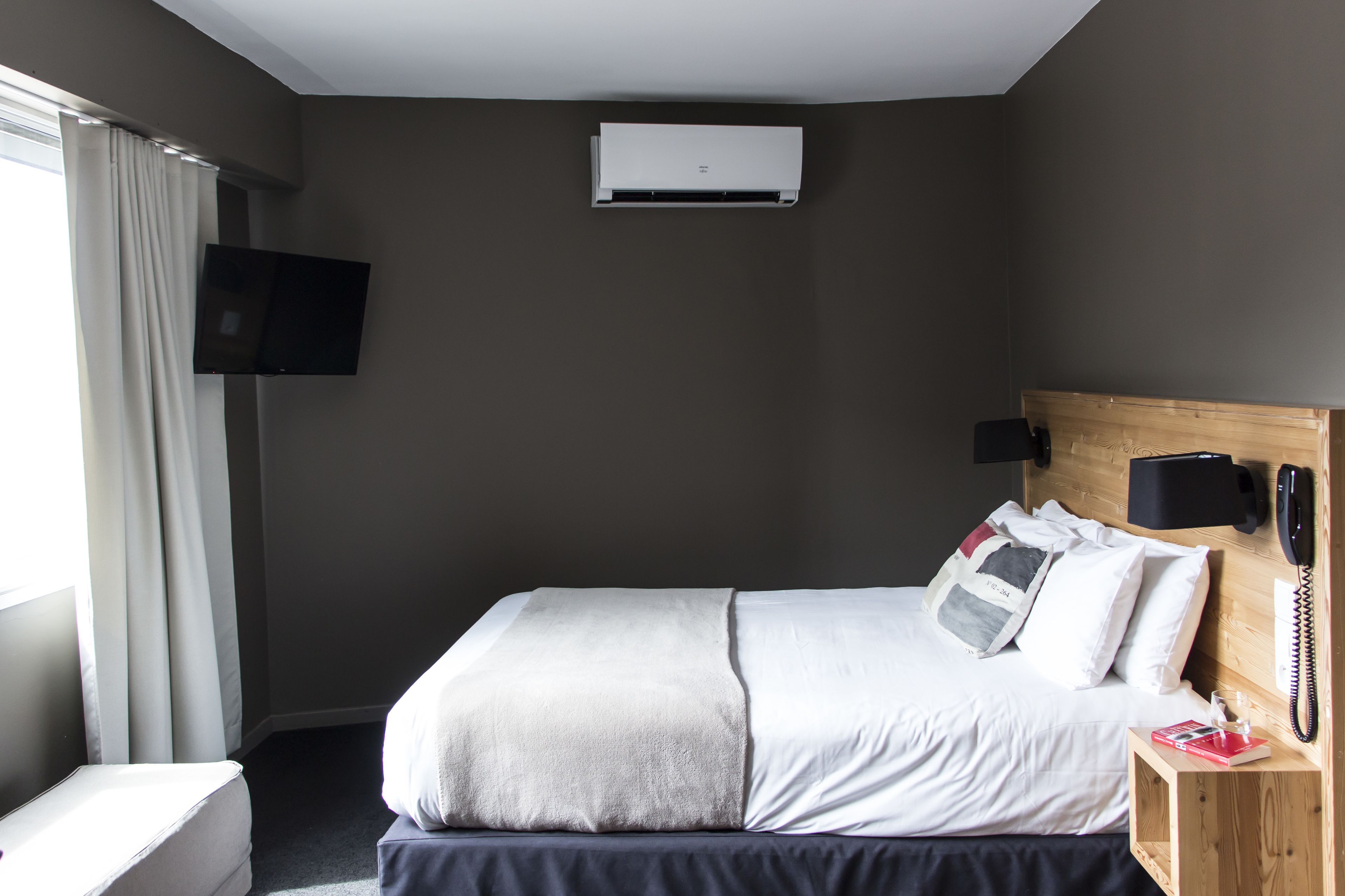 A hotel room with a bed, a TV mounted on the wall, an air conditioning unit, a bedside table with a phone, and a window with curtains.