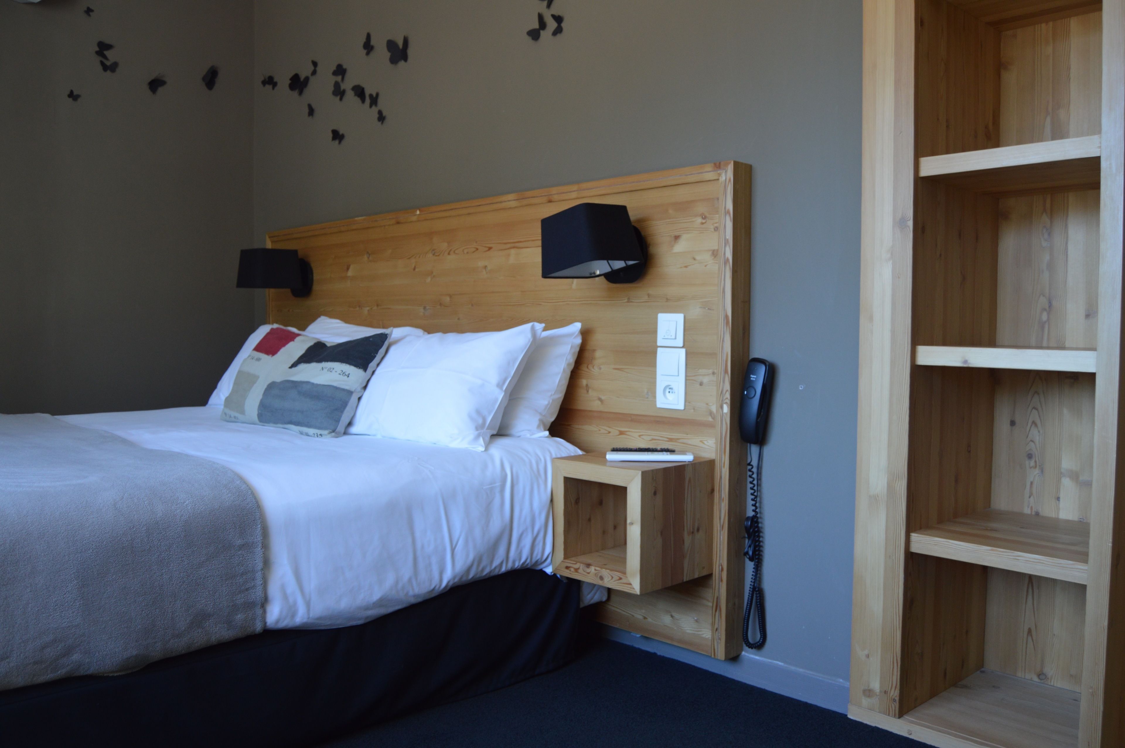 A neatly made bed with white linens and a black blanket, a wooden headboard, a bedside table with a lamp and a phone, and a wall with butterfly decals.