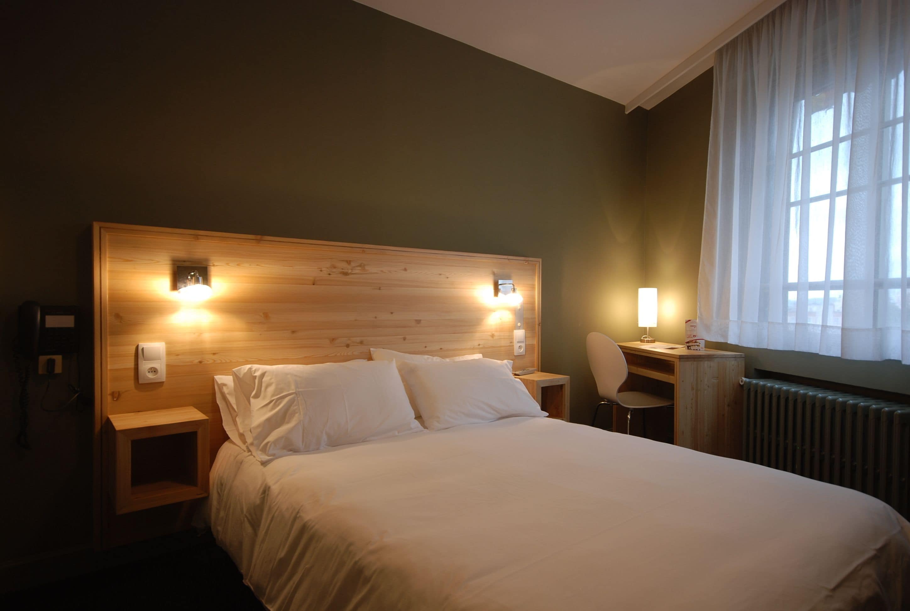 Ein Hotelzimmer mit einem hölzernen Kopfteil, zwei Nachttischlampen, einem Schreibtisch mit Stuhl und Blick ins Freie durch ein Fenster mit transparenten Vorhängen.