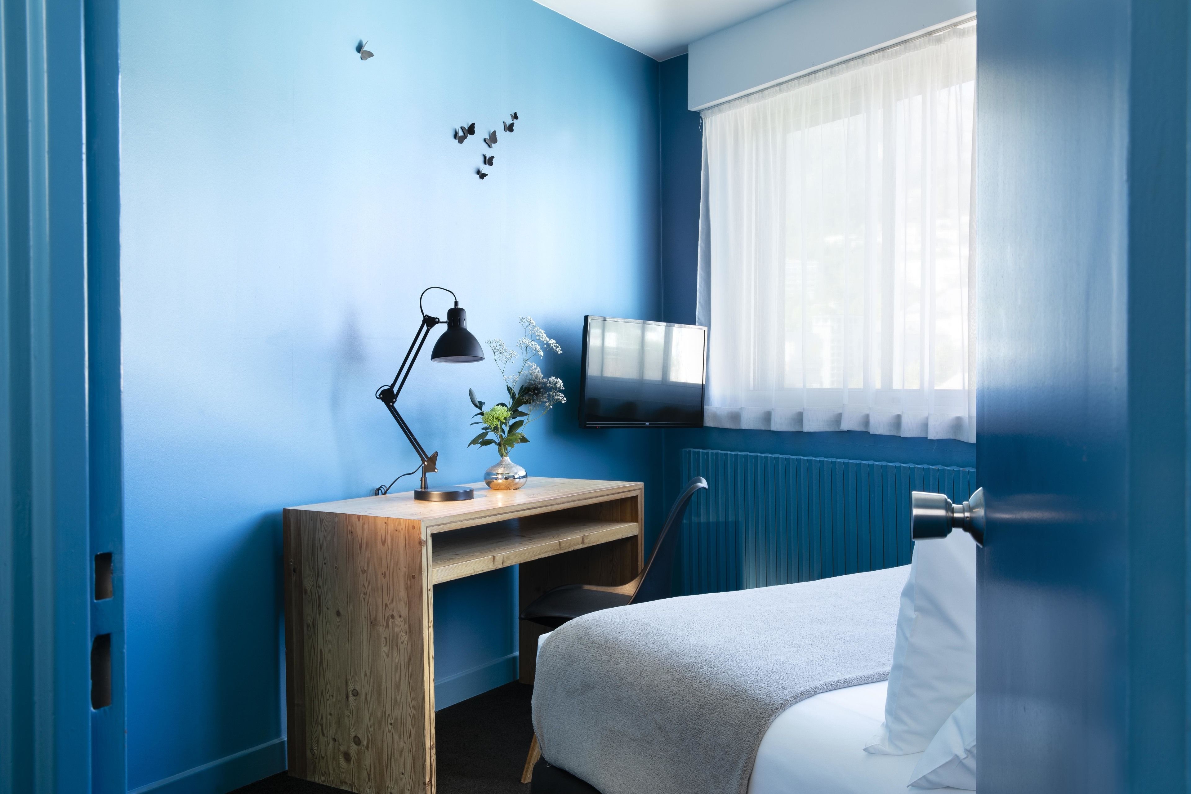 A bedroom with a bed, a desk, a chair, a lamp, and a TV on the wall.