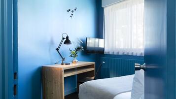 A bedroom with a bed, a desk, a chair, a lamp, and a TV on the wall.