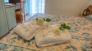 Un lit bien fait, avec une literie à motifs floraux et deux serviettes ornées de roses, dans une pièce comprenant un bureau et une chaise.