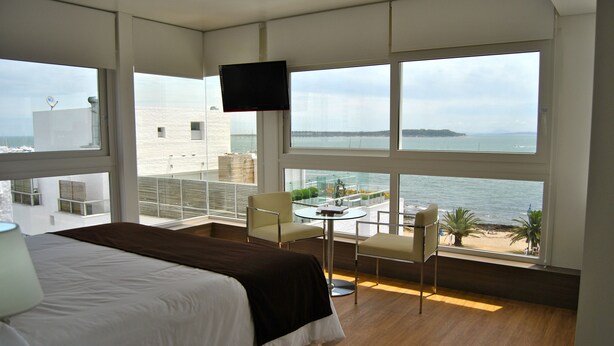 Ein Hotelzimmer mit einem großen Bett, einem an der Wand befestigten Fernseher, einem Balkon mit Tisch und Stühlen und Meerblick.