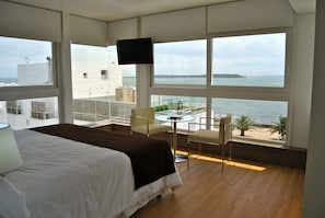 Ein Hotelzimmer mit einem großen Bett, einem an der Wand befestigten Fernseher, einem Balkon mit Tisch und Stühlen und Meerblick.