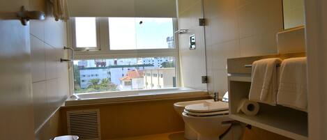 Un baño moderno con inodoro, lavamanos y ducha. Cuenta con un ventanal con vistas a edificios.