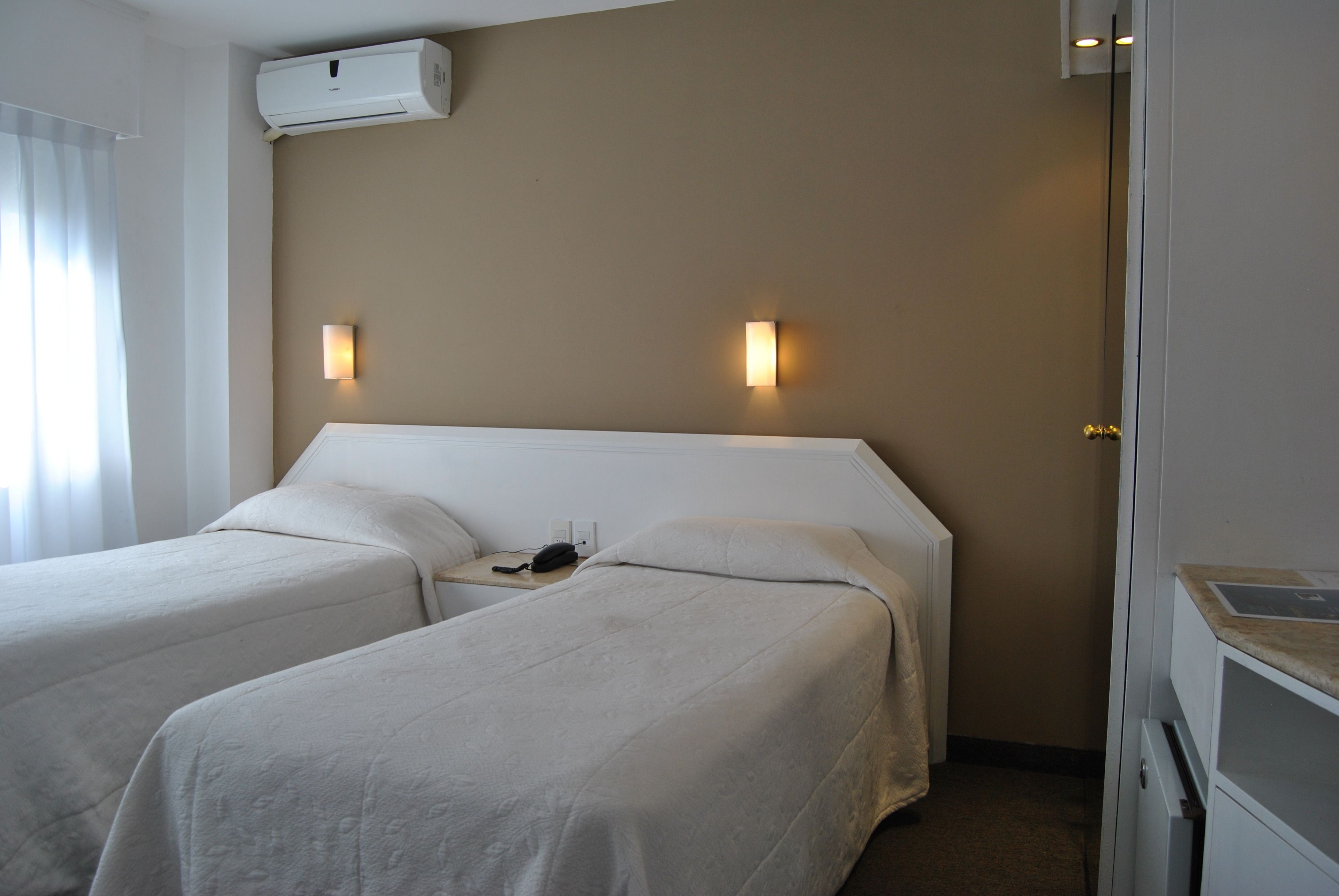 A hotel room with two single beds, a bedside table with a phone, and a wall-mounted air conditioner.