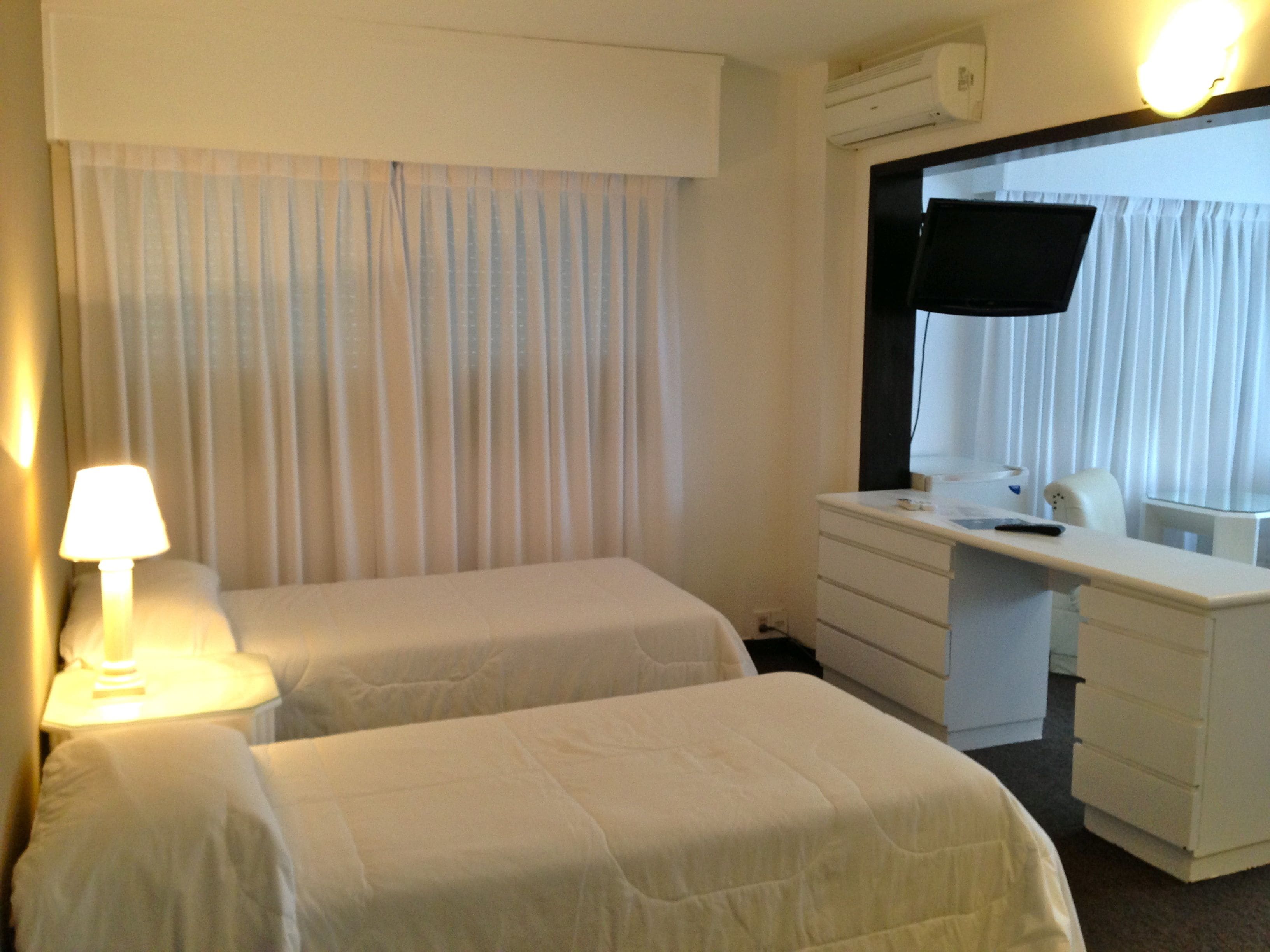A hotel room with two beds, a desk, a chair, a TV, and a lamp.