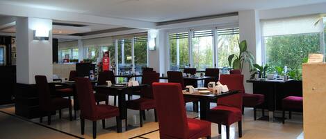 A spacious dining area with multiple tables and chairs, large windows offering a view of greenery, and a clean, modern interior design.