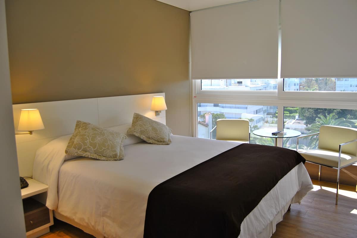 Habitación de hotel con una cama grande, dos lámparas de noche, una mesita con una silla y vistas a la ciudad a través de amplios ventanales.