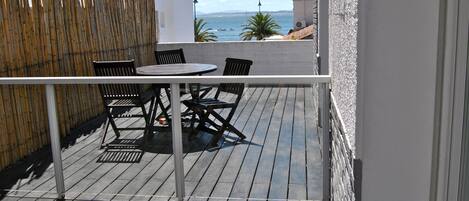 Una terraza de madera con una mesa redonda y cuatro sillas, con vista a la costa y el mar, donde se ven embarcaciones.