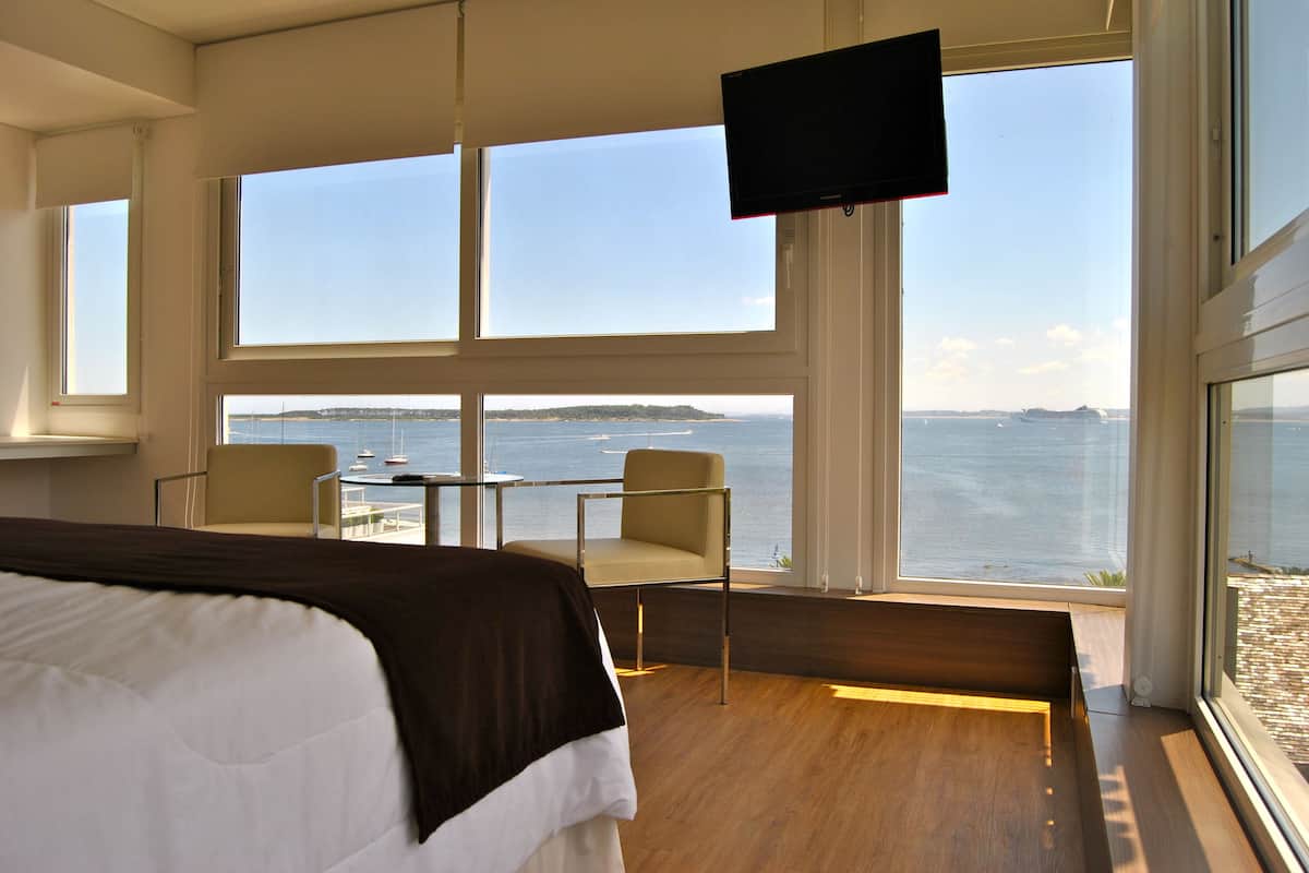 Habitación de hotel con una cama grande, televisión, balcón con vistas al mar y una mesa con sillas.