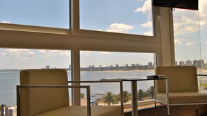 Ein Zimmer mit großem Fenster mit Blick auf Strand und Stadtpanorama. Zwei Stühle und ein kleiner Tisch sind vorhanden.