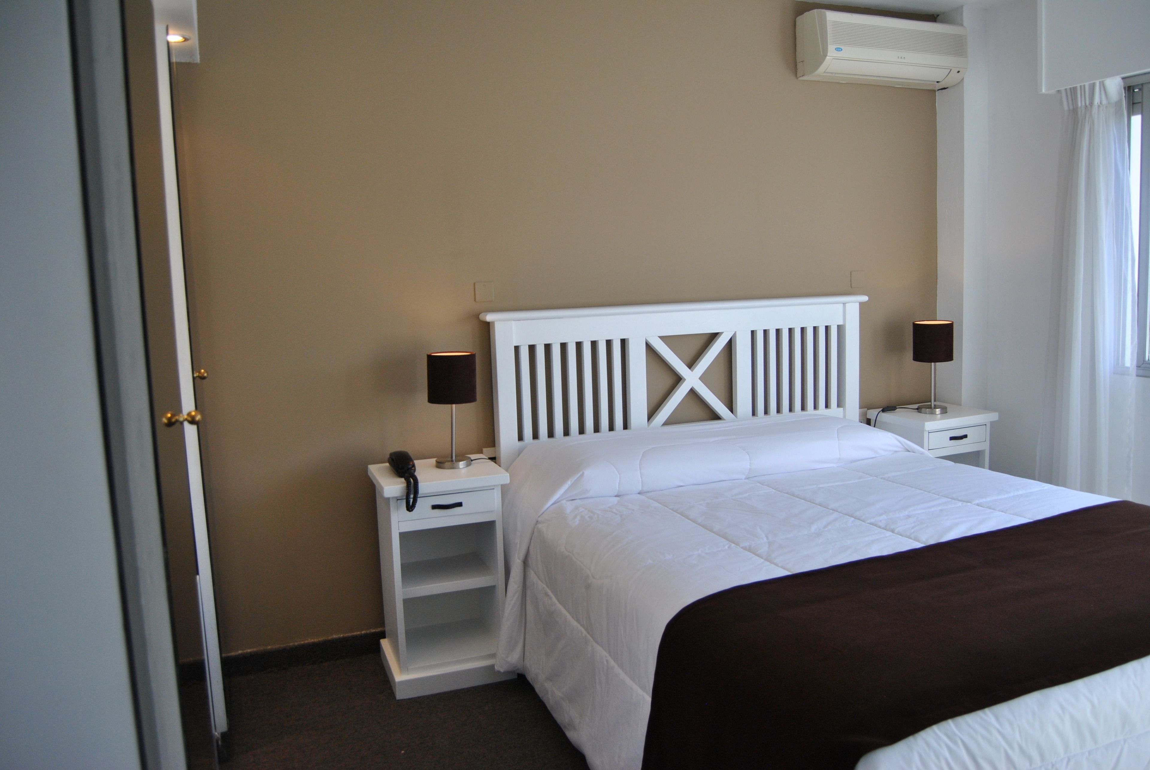 A hotel room with a white bed, two bedside tables, a white nightstand, a telephone, and a wall-mounted air conditioner.