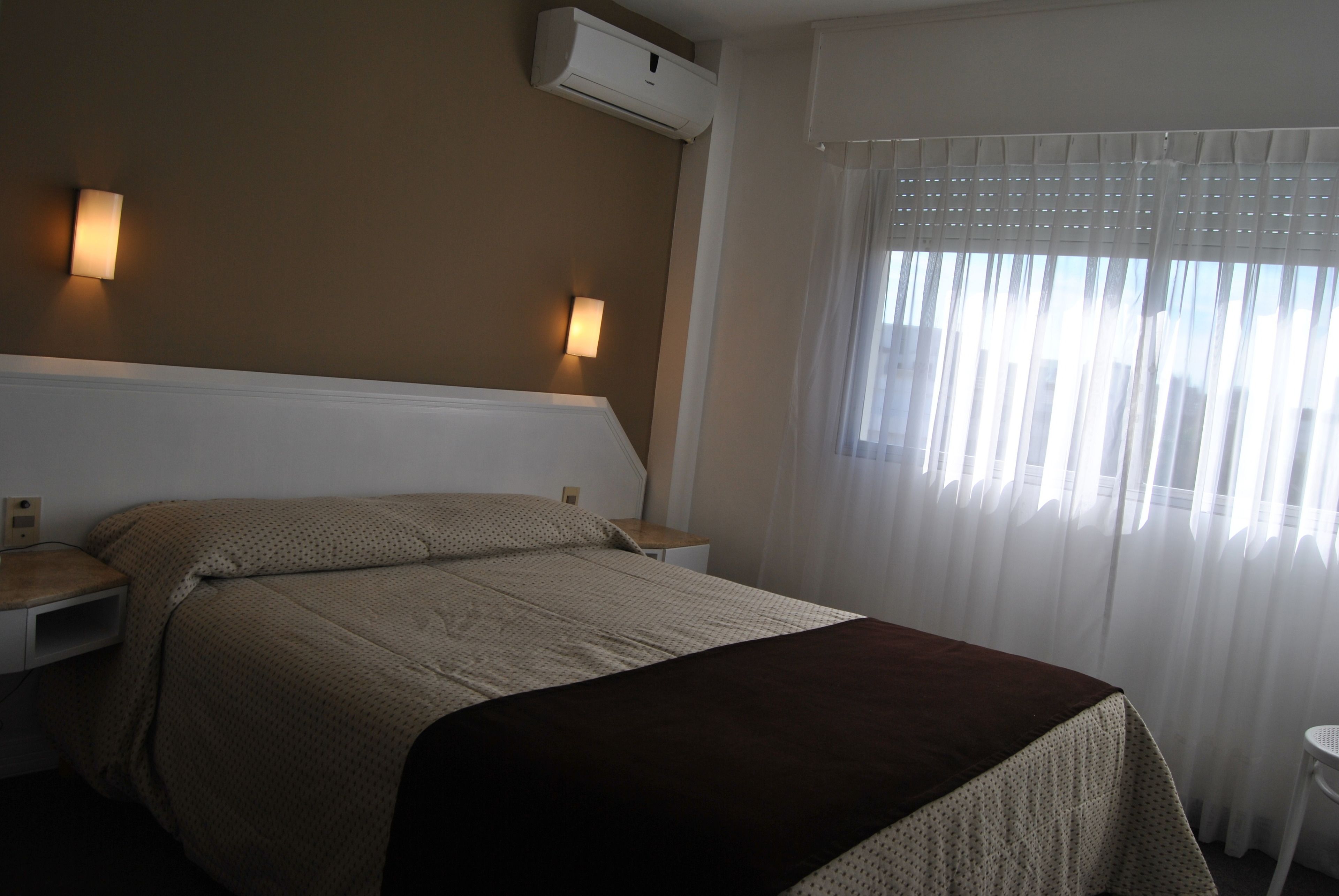 Una habitación de hotel con una cama, una mesita de noche, aire acondicionado de pared y una ventana con cortinas translúcidas.