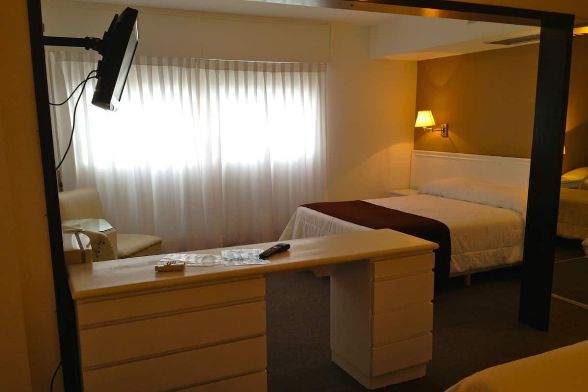 Habitación de hotel con cama, escritorio con cajones, silla, lámpara y ventana con cortinas.