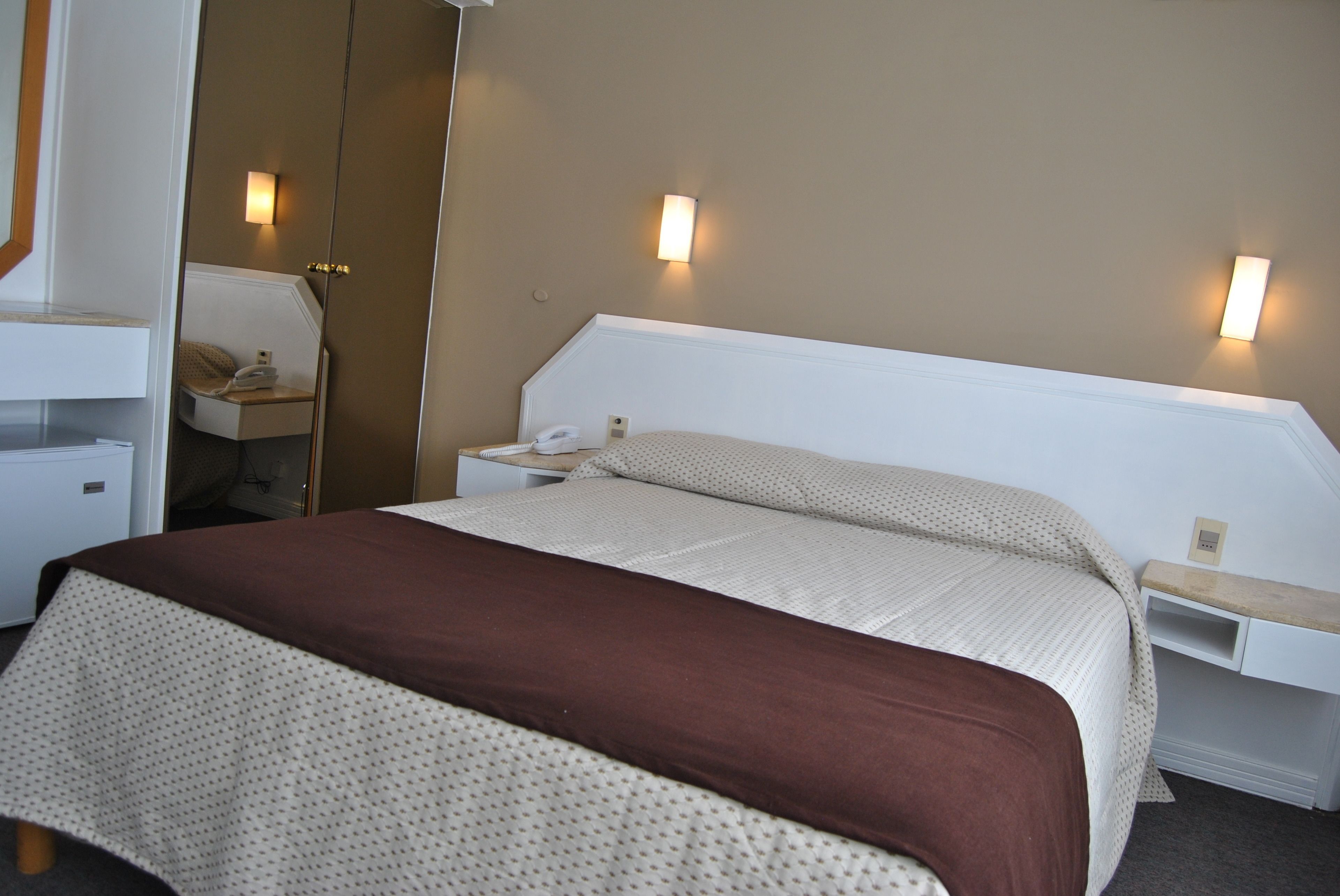 Une chambre d’hôtel bien aménagée, avec un grand lit, des tables de chevet, un miroir et deux lampes fixées au mur.