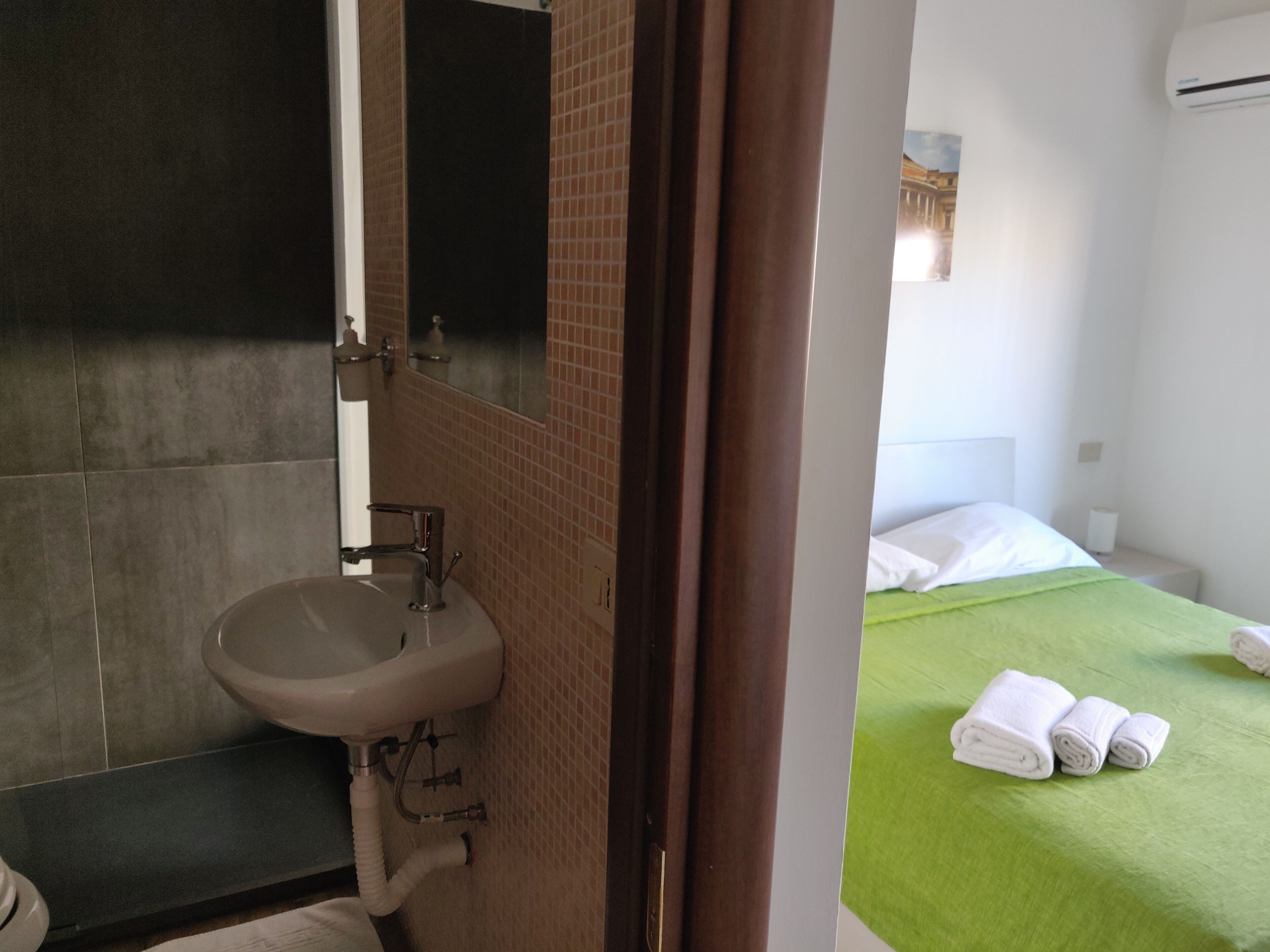 A bathroom with a sink, mirror, and towel rack. There is a bed with a green bedspread and rolled towels.