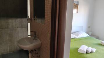A bathroom with a sink, mirror, and towel rack. There is a bed with a green bedspread and rolled towels.