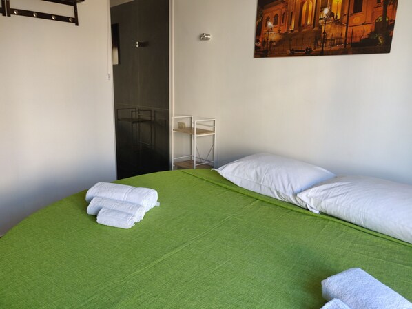A single bed with a green bedspread and white pillows, a white wall, and a painting of a building.
