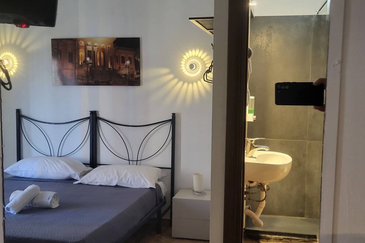 Un dormitorio con cama, mesita de noche, un cuadro en la pared y un baño con lavamanos y ducha.