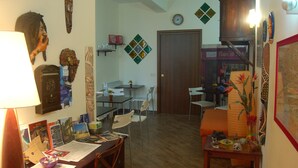 Una habitación con piso de madera, un escritorio con papeles y una lámpara, una silla, una puerta y adornos en la pared que incluyen máscaras y un reloj.