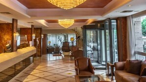 A hotel lobby with a large chandelier, leather seating, a reception desk, and a view through large glass doors. - Park (Volos)