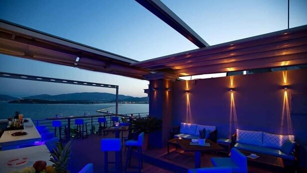 A deck with seating and a table, overlooking a body of water and hills. - Park (Volos)