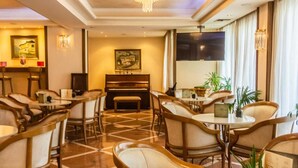 A hotel lobby with round tables and chairs, a piano, and a large painting on the wall. - Park (Volos)