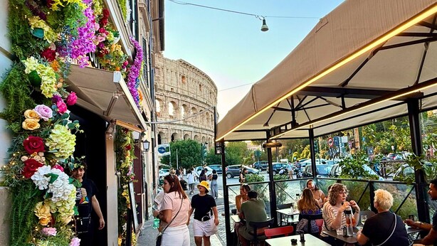 An outdoor seating area with tables and chairs, a view of the Colosseum in the background, and a decorative floral arrangement on the left. - First Floor (Rome)