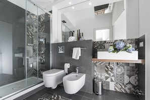 Features a sleek glass-enclosed shower, white toilet, and bidet, with geometric patterned tiles and a large mirror.