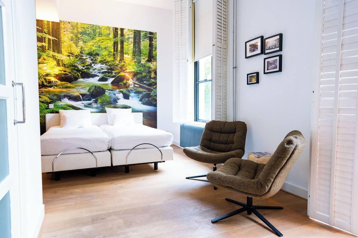 A room with a bed, two armchairs, a large forest mural, and a window with blinds.