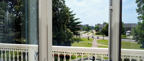 A balcony with ornate white railings, a view of a landscaped garden with a fountain, and a road with vehicles and buildings in the background.