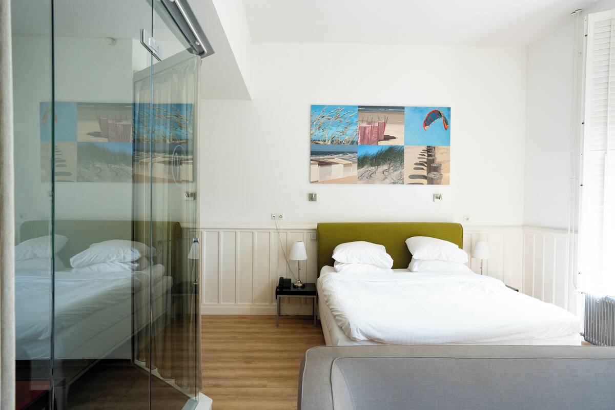 A hotel room with a bed, a nightstand, a glass partition, and a wall with a beach-themed artwork.