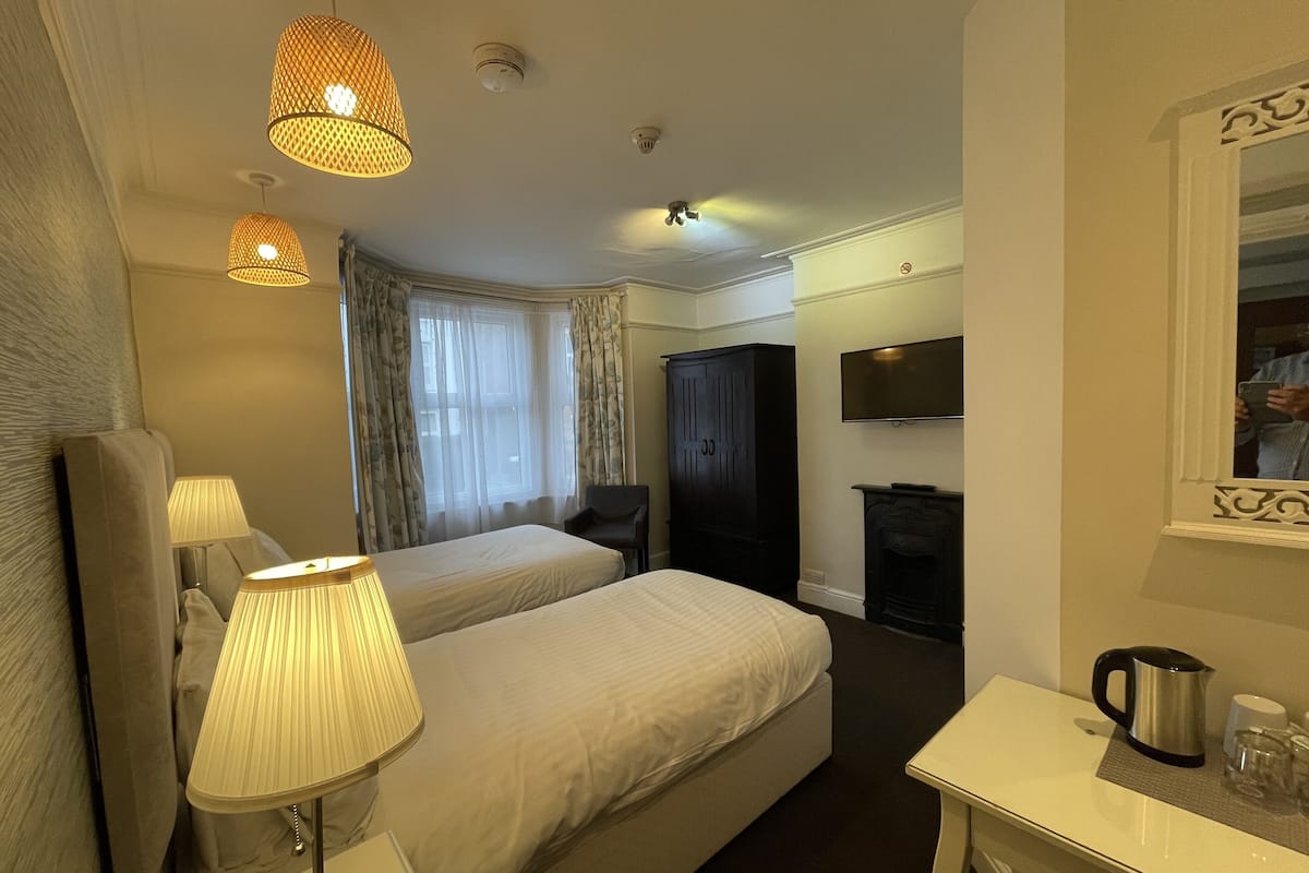 A hotel room with a bed, bedside lamps, a desk with a kettle, a television, and a fireplace.