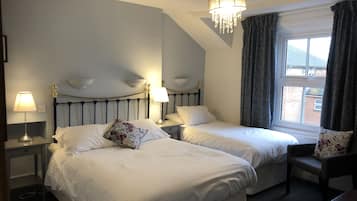 A hotel room with two beds, a chandelier, a window with curtains, and a desk with a lamp.