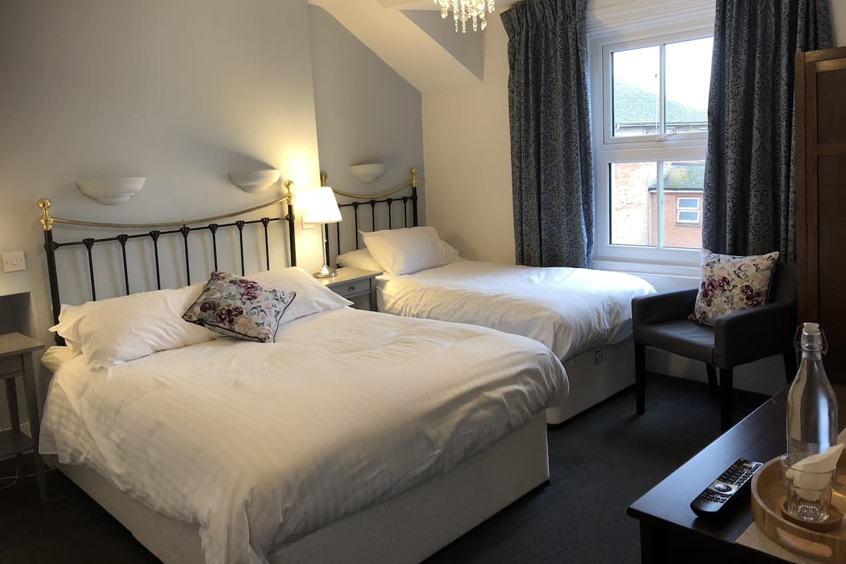 A hotel room with two beds, a nightstand with a lamp, a chair, a window with curtains, and a table with a remote control and a glass.