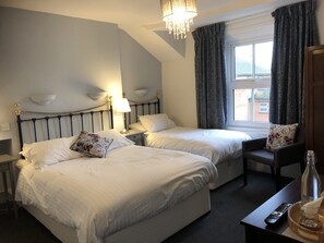 A hotel room with two beds, a nightstand with a lamp, a chair, a window with curtains, and a table with a remote control and a glass. - Newton House (Oxford)