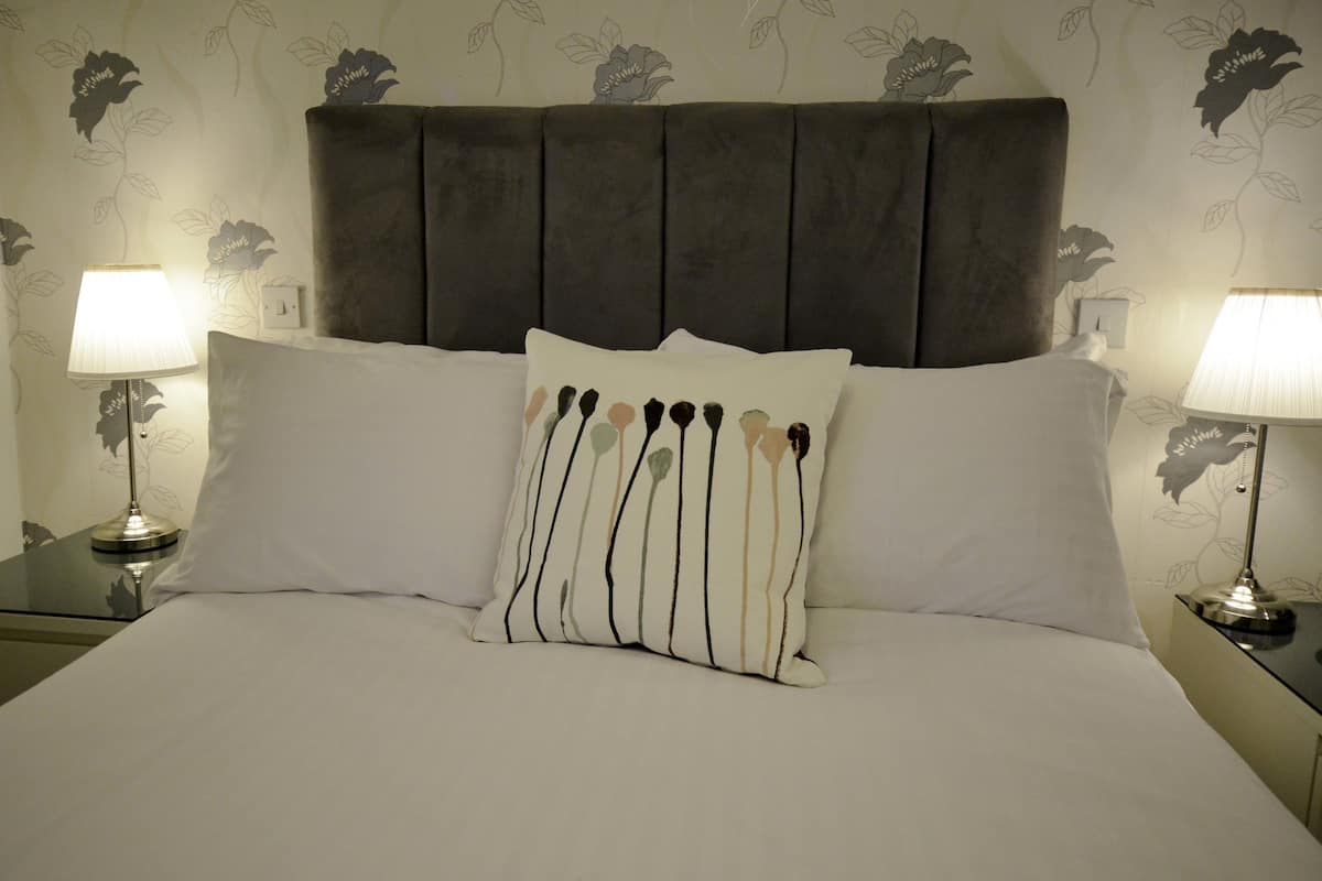 A neatly made bed with a grey upholstered headboard, two bedside lamps, and a floral-patterned wall.