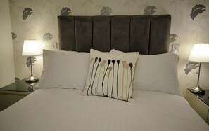A neatly made bed with a grey upholstered headboard, two bedside lamps, and a floral-patterned wall. - Newton House (Oxford)