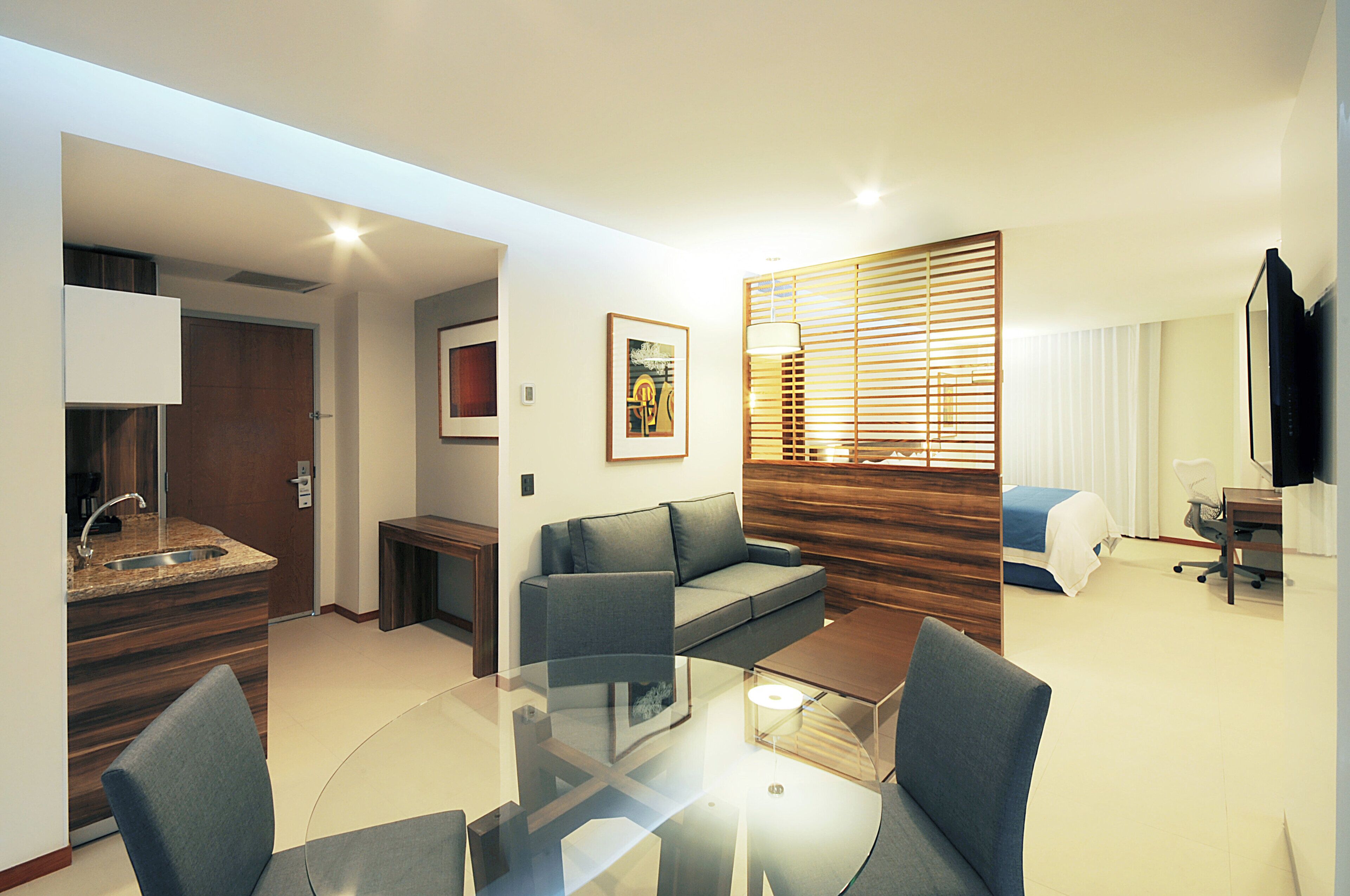 A modern hotel room with a kitchenette, a sofa, a glass coffee table, and a bed.