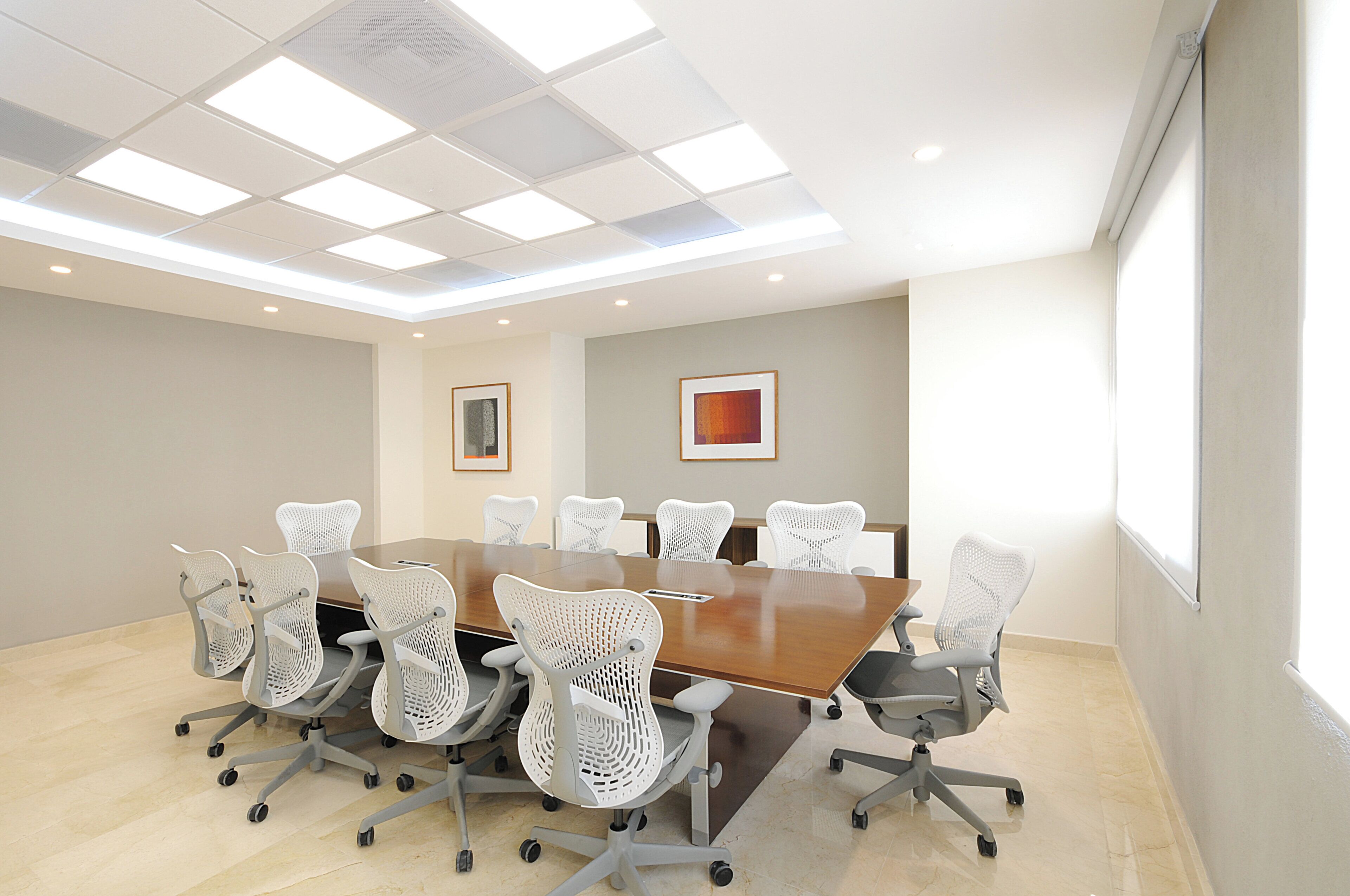 A conference room with a large table surrounded by white chairs, a ceiling with square lights, and two framed pictures on the walls.