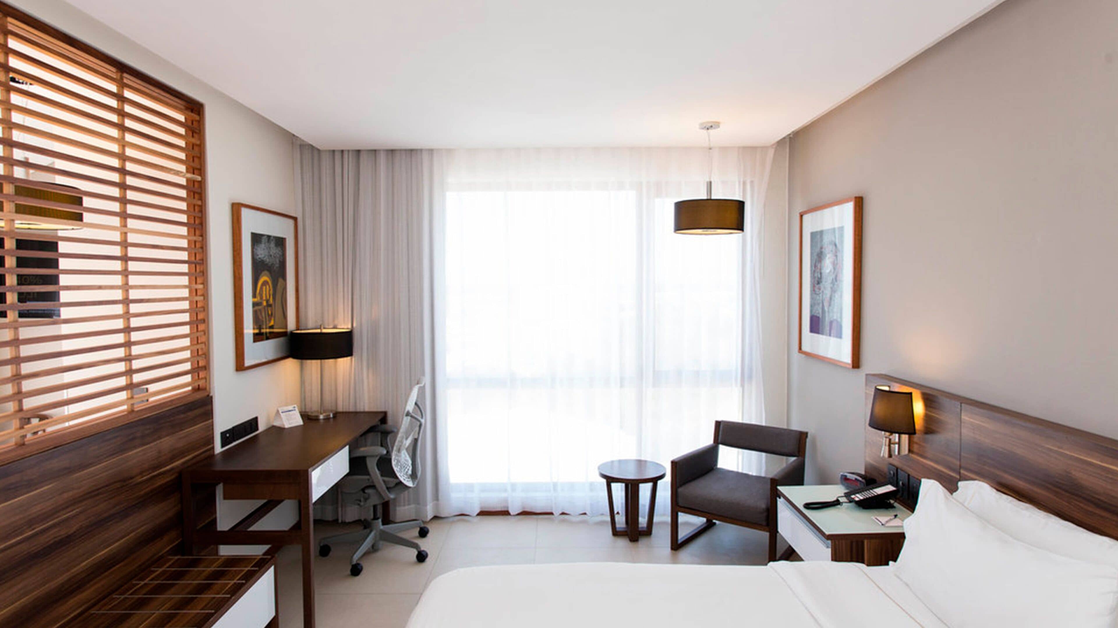 A hotel room with a bed, desk, chair, and a small table. There are two windows with blinds, a ceiling light, and two wall-mounted lamps.
