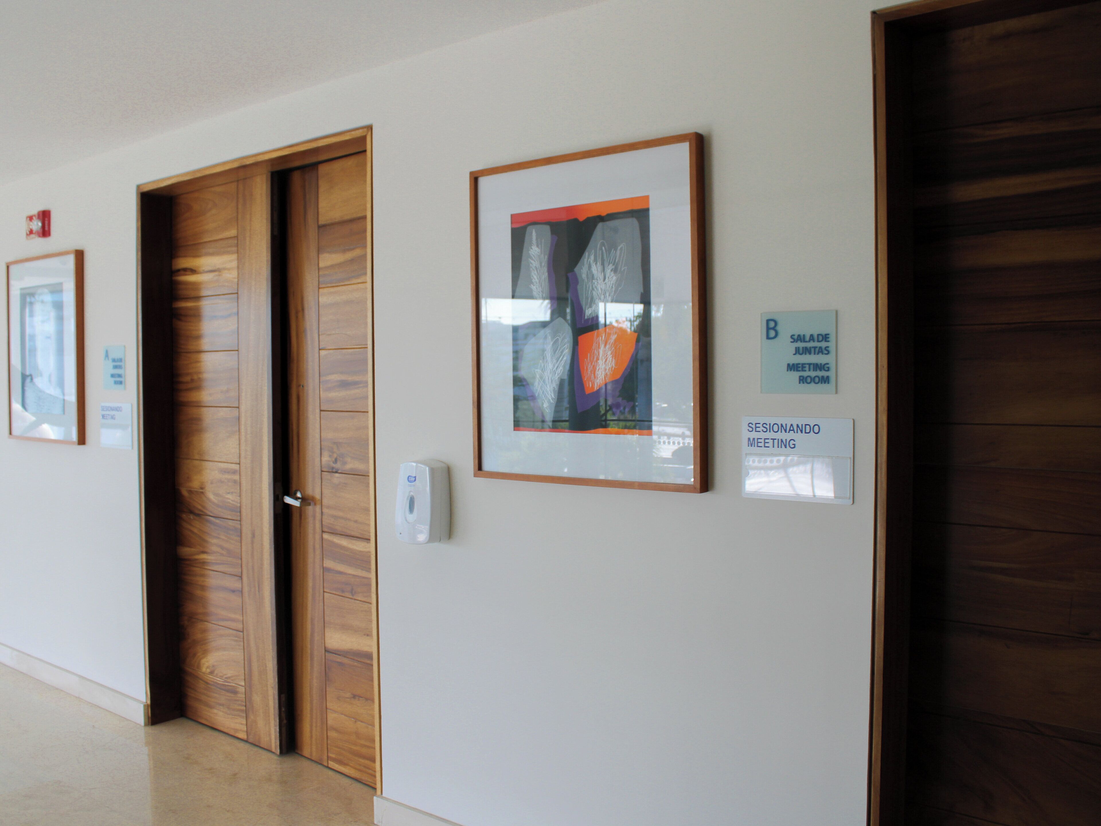 Un pasillo con puertas de madera, obras de arte enmarcadas en las paredes y señalizaciones que indican nombres de habitaciones y lugares de reuniones.