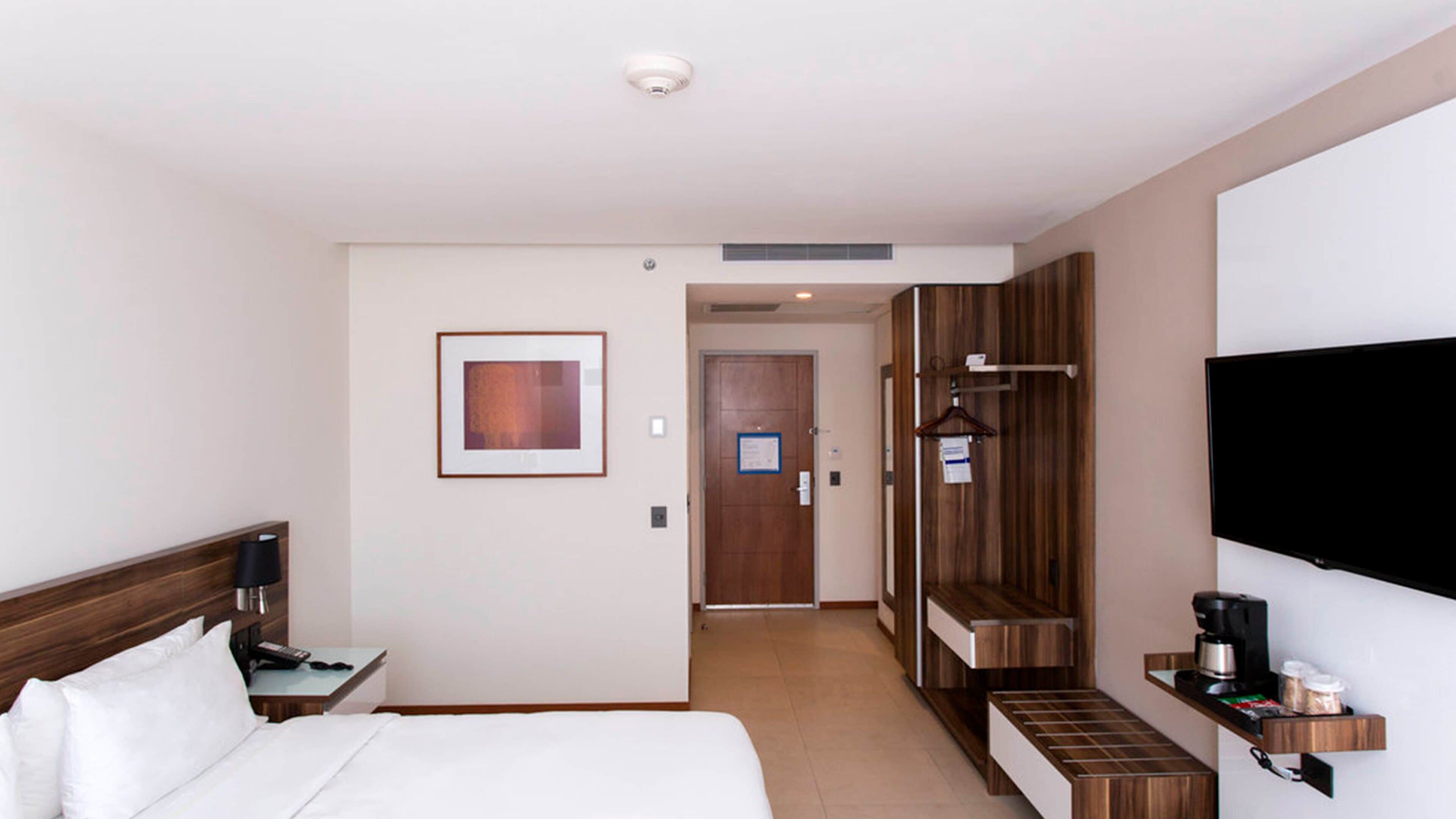 Une chambre d’hôtel comprenant un lit, un bureau avec une lampe, un téléviseur fixé au mur et une armoire en bois.